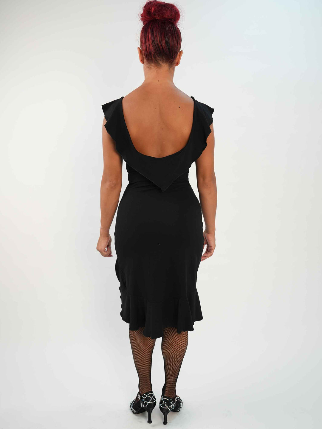 Femme portant une robe noire de danse latine sans manches, avec encolure volantée et ourlet asymétrique, associée à des collants résille et des chaussures de danse ornées. Tenue élégante et fluide, parfaite pour la salsa ou le ballroom. - vue de l'arrière
